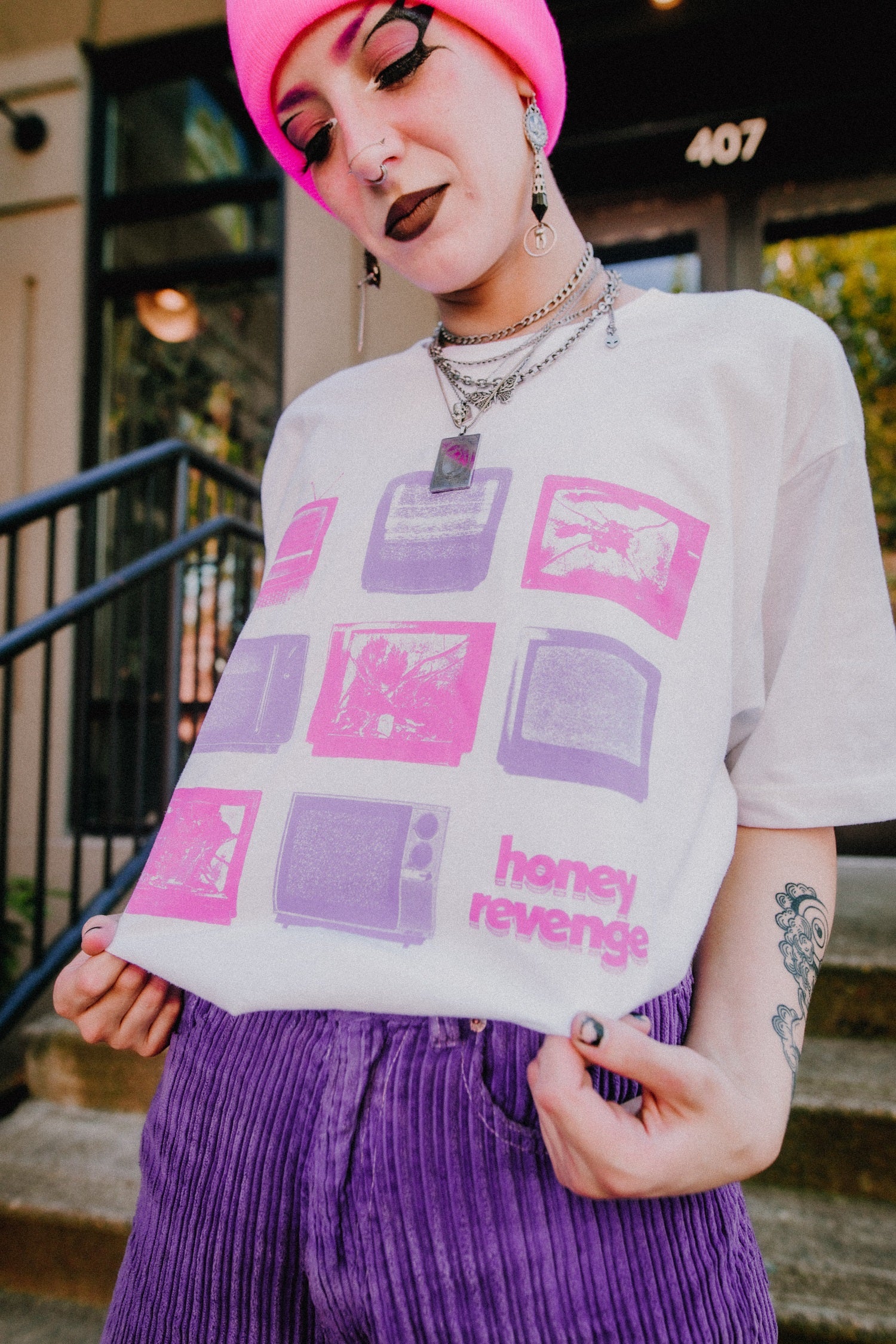 Wearing a neon pink beanie, layered necklaces, and a white Honey Revenge T-shirt with purple and pink graphics, a person stands outside by stairs, showing off tattoos on their arm and their bold style.