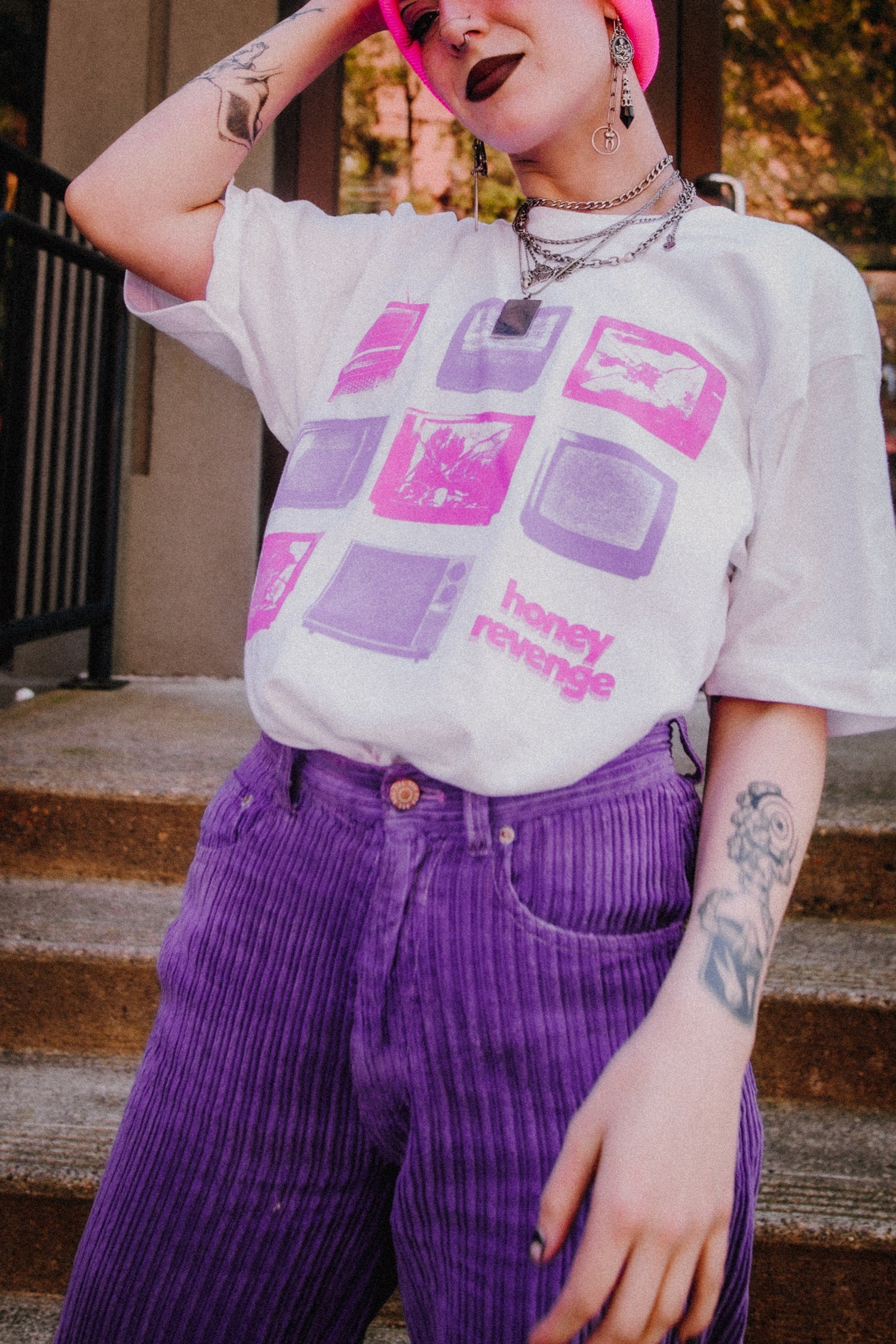 A person wearing a Honey Revenge white graphic t-shirt, pink beanie, purple pants, and layered necklaces stands on outdoor steps, displaying tattoos on their arm and hand.